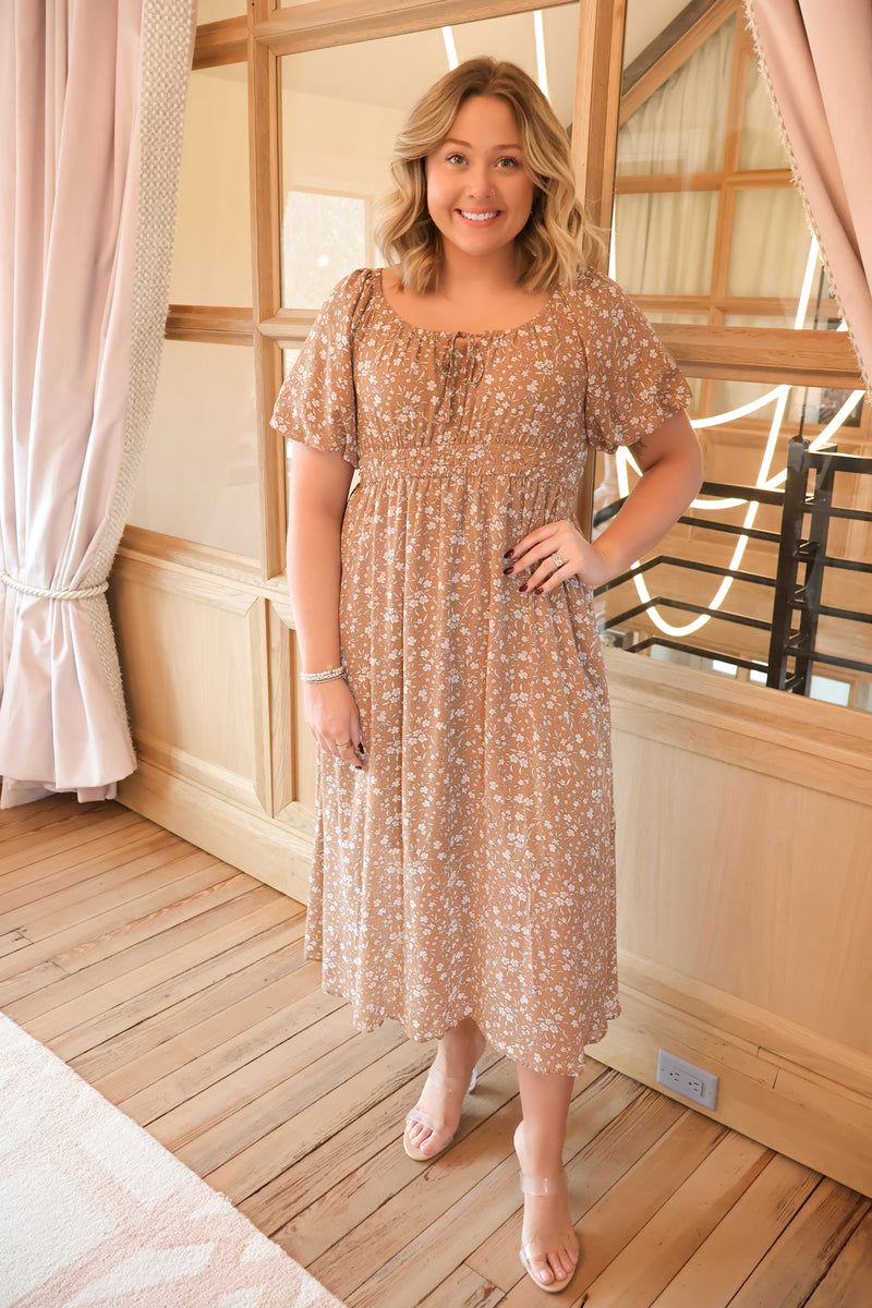 Model wearing a mocha floral midi dress featuring a front tie, smocked waist, side pockets, and a comfortable fully lined design, perfect for any occasion.